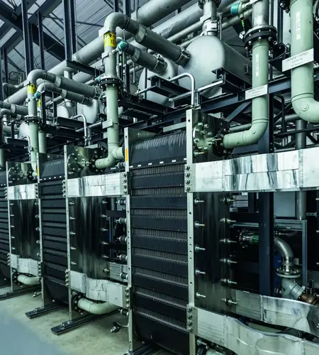 Industrial interior view focusing on large metallic machinery with an array of pipes, valves, and metal structures.